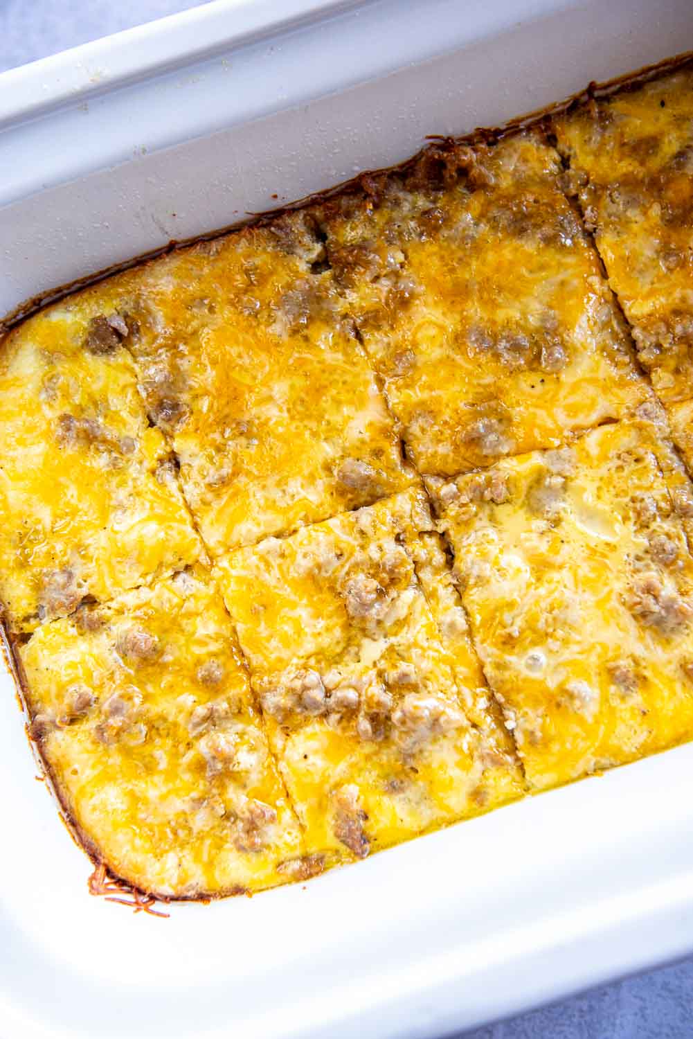 White baking dish with crescent rolls dough laid out and beaten eggs poured on top with ground pork breakfast sausage on top and shredded cheese on top cooked cut into 8 squares