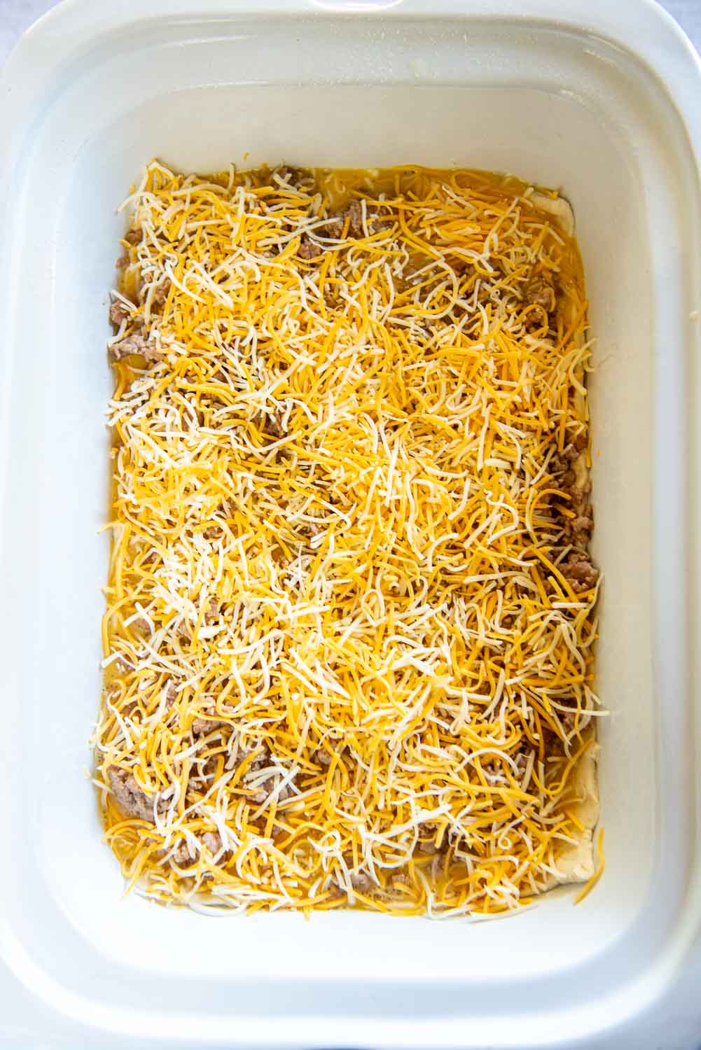 White baking dish with crescent rolls dough laid out and beaten eggs poured on top with ground pork breakfast sausage on top and shredded cheese on top