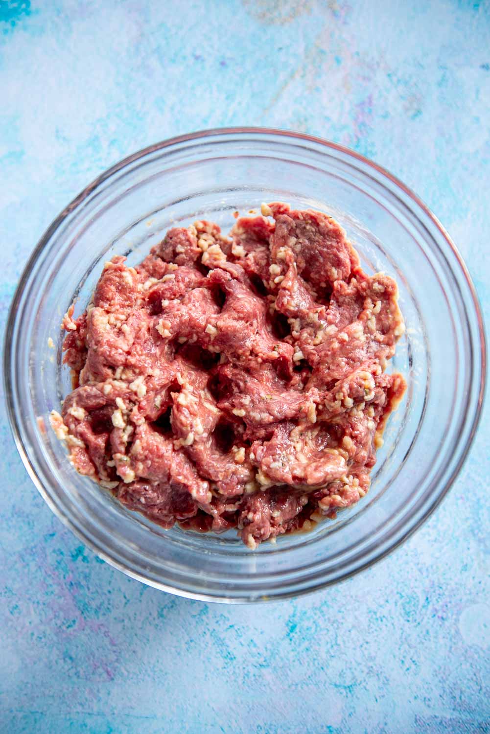 Raw ground beef covered in seasoning in a clear bowl