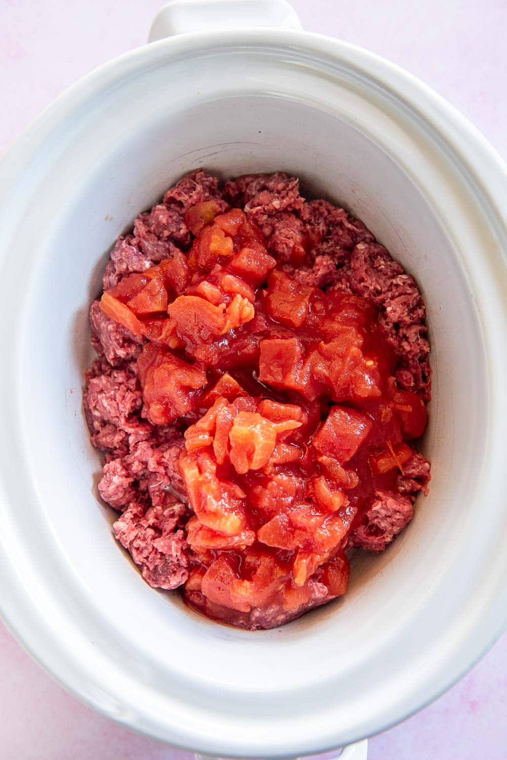 raw ground beef broken apart with tomatoes in sauce on top inside of white slow cooker bowl