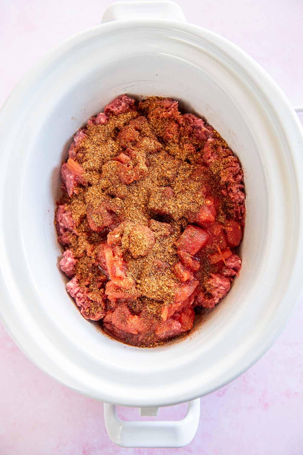 raw ground beef with seasonings and red cause inside of a white slow cooker