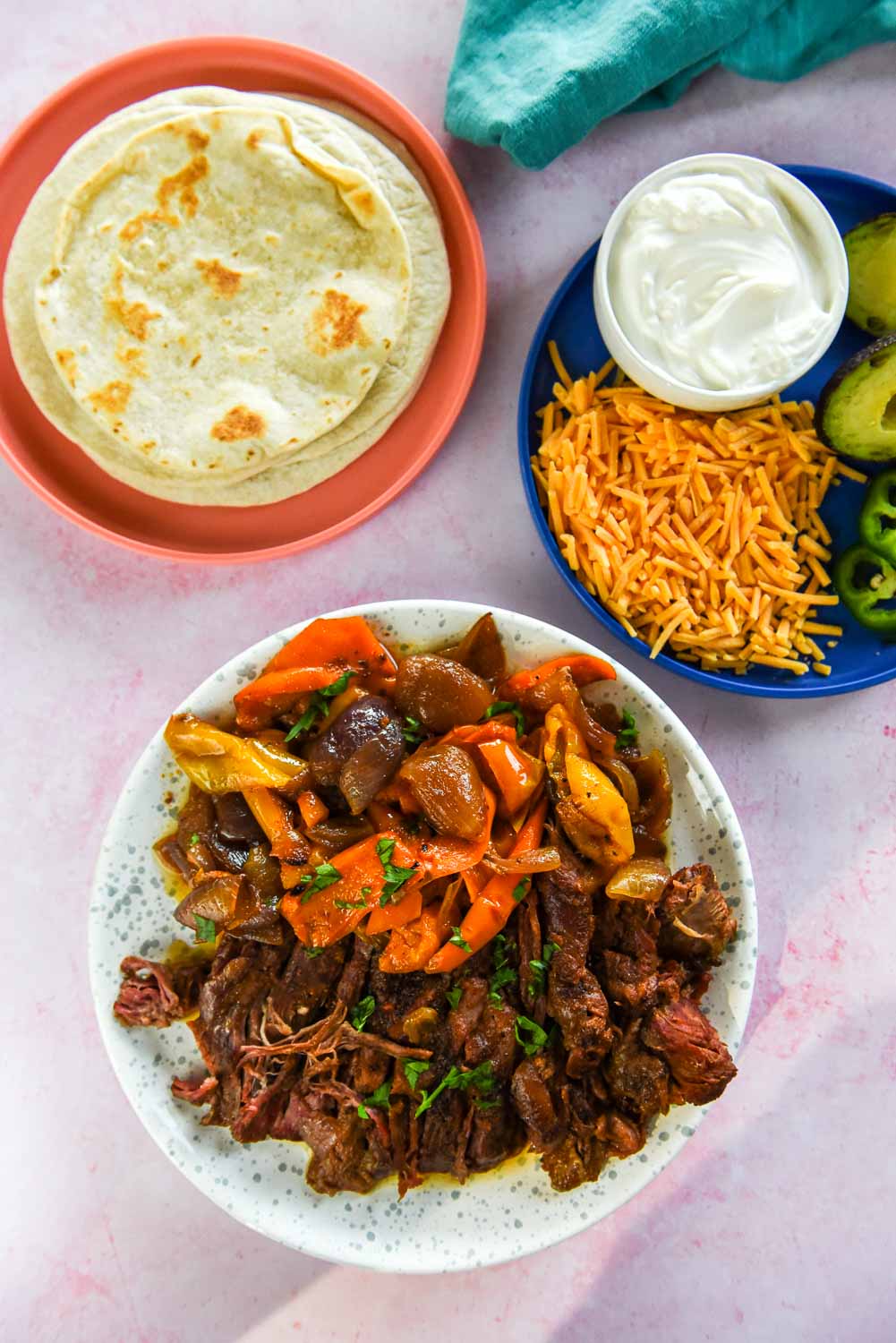 white with blue speckles plate with sliced fajita steak and veggies next to blue plate with cheese and sour cream and coral plate with tortillas