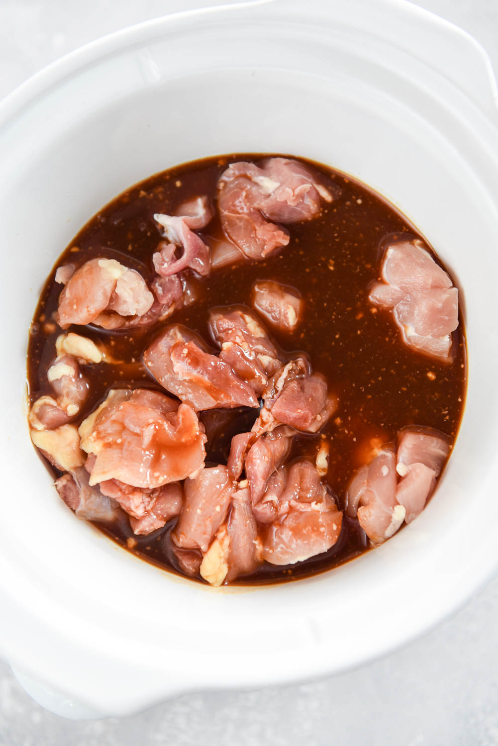 Raw chicken inside of sauce mixture in white slow cooker bowl