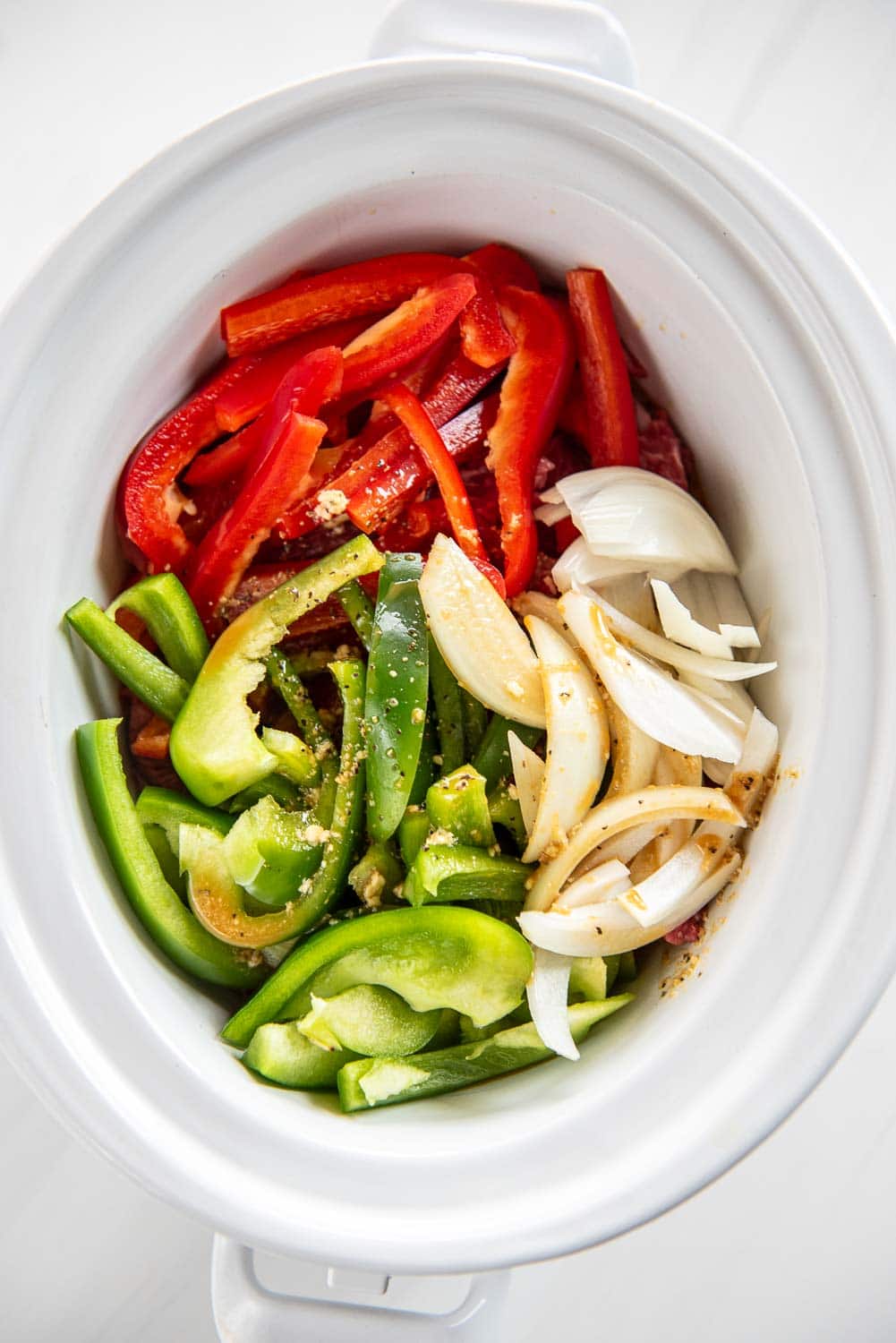 raw green peppers, red peppers, onions, all sliced inside of slow cooker with seasoning on top