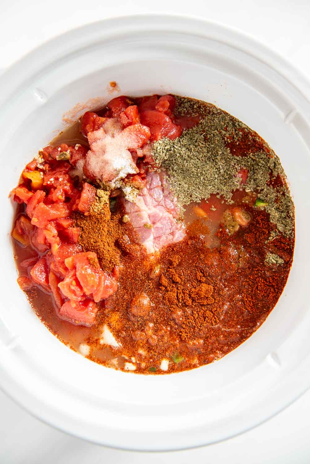 raw pork shoulder roast cut with lines with chopped onions, green peppers, minced garlic on top, tomato sauce and diced tomatoes, and seasonings on top inside of slow cooker