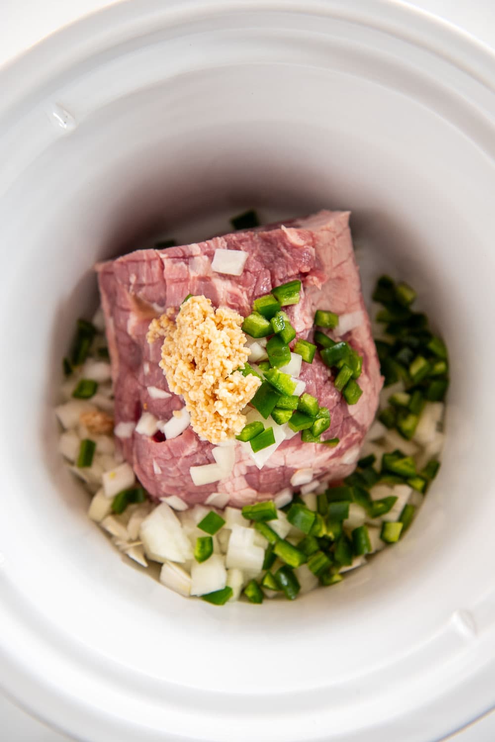 raw pork shoulder roast cut with lines on top inside of slow cooker with chopped onions, green peppers, and minced garlic on top