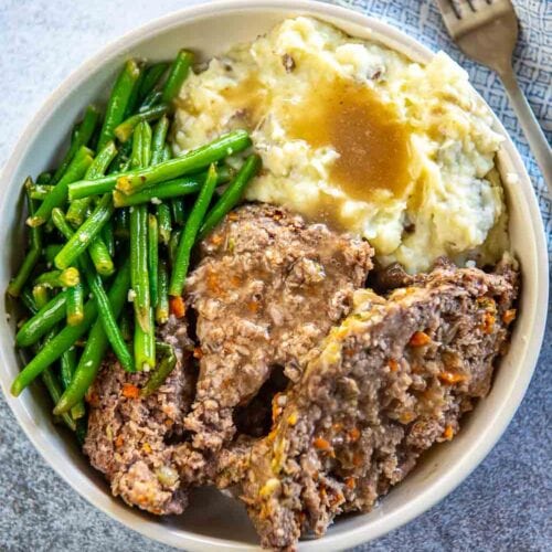 two slices of meatloaf with mashed potatoes, green beans and gravy in a shallow bowl