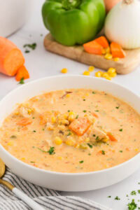 White shallow bowl of chowder with corn and sweet potatoes with green pepper, onion and sweet potato in background