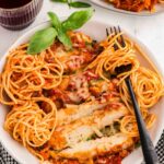 plate of sliced chicken parmesan with swirls of pasta noodles in a white bowl with a black fork next to a glass of red wine