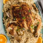A large serving dish featuring a bed of cooked rice, topped with a whole cooked chicken and elegantly garnished with fresh orange slices.