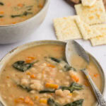 A bowl of the cooked bean soup, served with a spoon laid on top, showcasing the white navy beans, carrots, spinach, and creamy broth. The bowl is ready to be enjoyed.