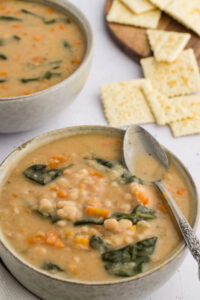 A bowl of the cooked bean soup, served with a spoon laid on top, showcasing the white navy beans, carrots, spinach, and creamy broth. The bowl is ready to be enjoyed.
