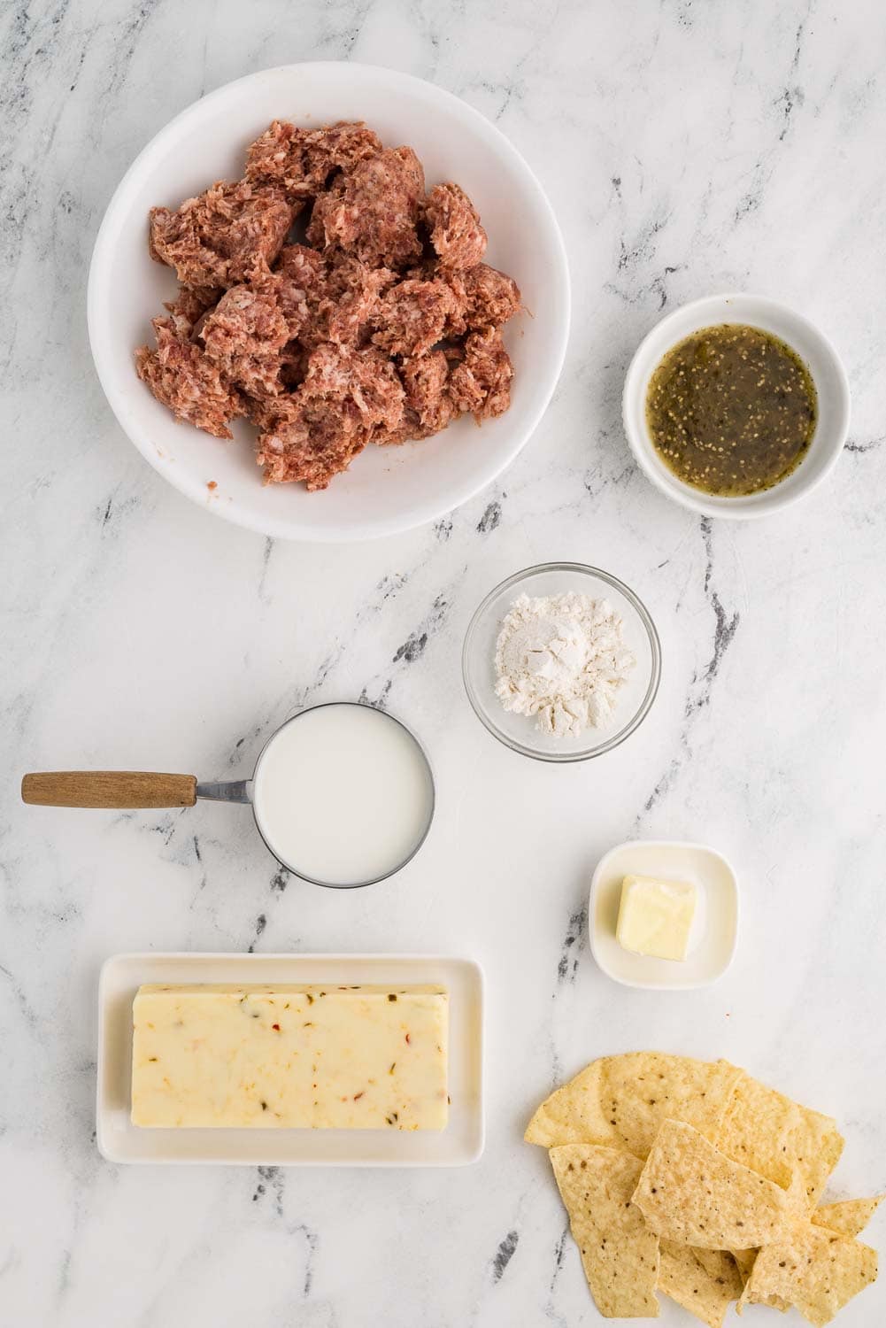 Ingredients for a sausage cheese dip recipe arranged on a kitchen counter. Includes 12 oz of ground pork sausage, an 8 oz block of pepper jack cheese, a small bowl with 2 tablespoons of flour, a stick of butter, a measuring cup with 1 cup of milk, a small bowl of salsa verde, and tortilla chips laid on counter