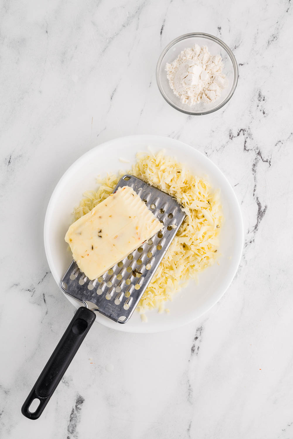 : A bowl filled with shredded pepper jack cheese, with a box grater laid across the top. A partially used block of pepper jack cheese rests on the grater, ready to be shredded further.