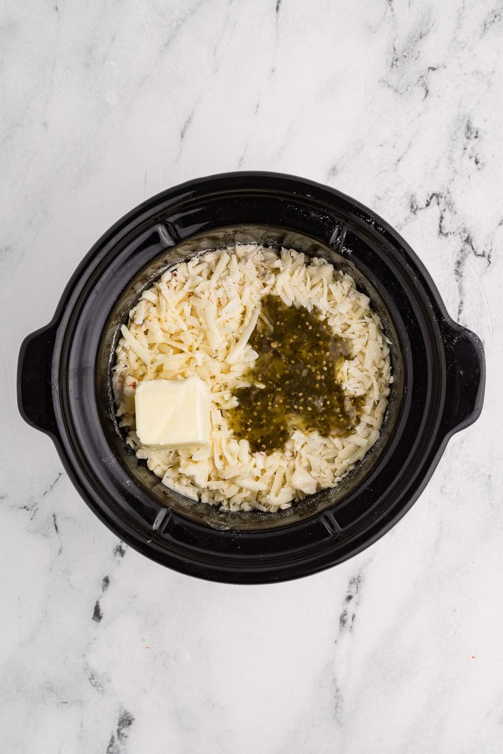 Ingredients for sausage queso dip inside a slow cooker. Includes cooked ground pork sausage, shredded pepper jack cheese, flour, butter, milk, and salsa verde, layered and ready to cook.