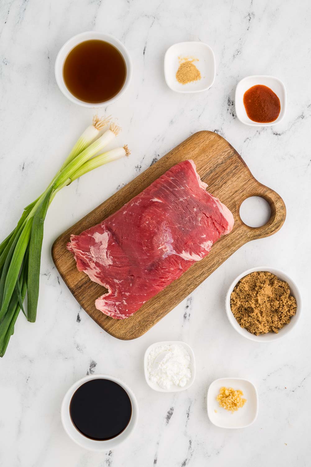 Ingredients for shredded Mongolian beef recipe on a counter: 1 1/2 pounds of flank steak, 1/4 cup soy sauce, 1/4 cup beef broth, 1/4 cup dark brown sugar, 1 teaspoon jarred minced garlic, 1/4 teaspoon powdered ginger, 1 teaspoon Sriracha sauce, 2 tablespoons corn starch, and 1/2 bunch green onions