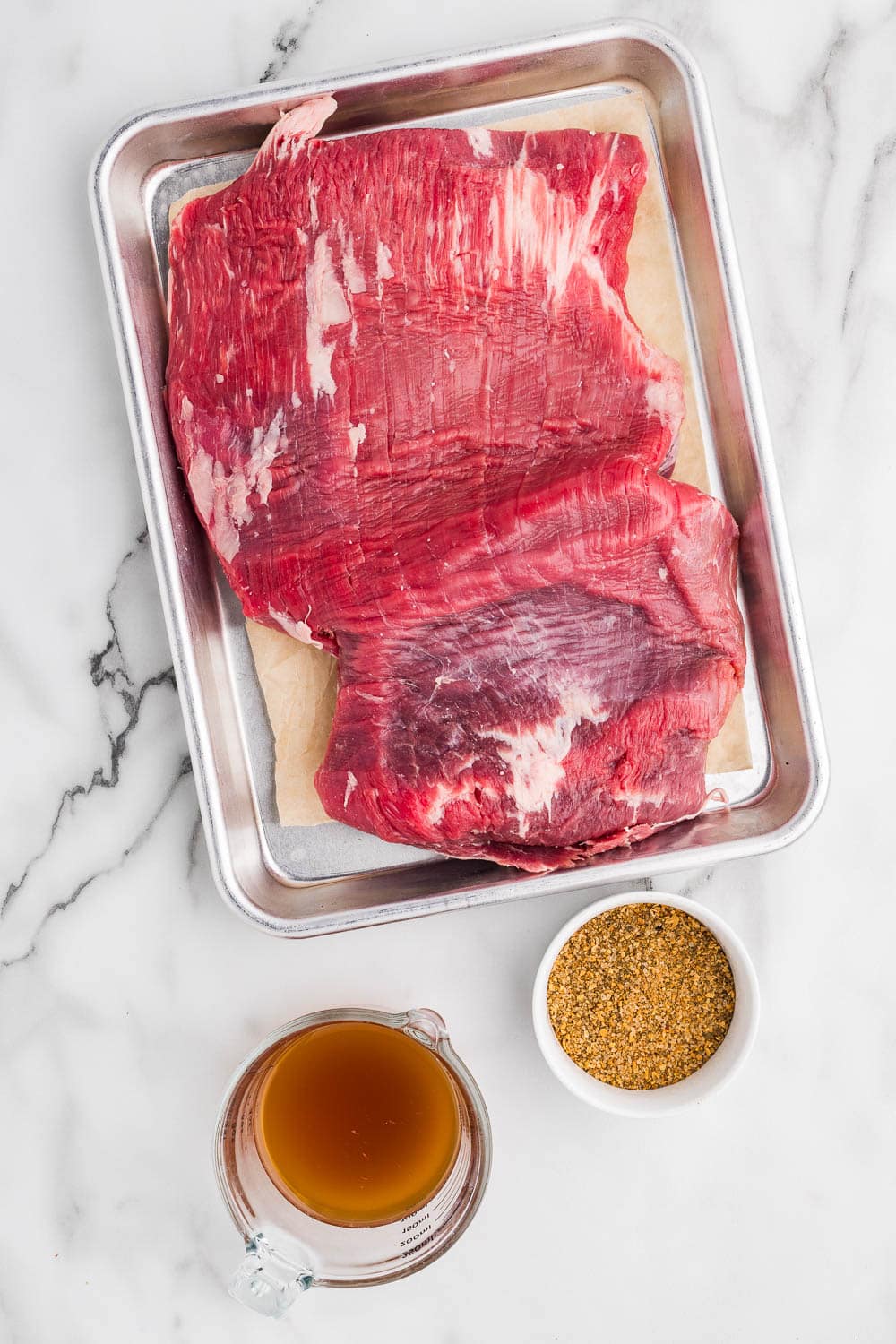 Raw flank steak on a baking sheet with spices and seasoning