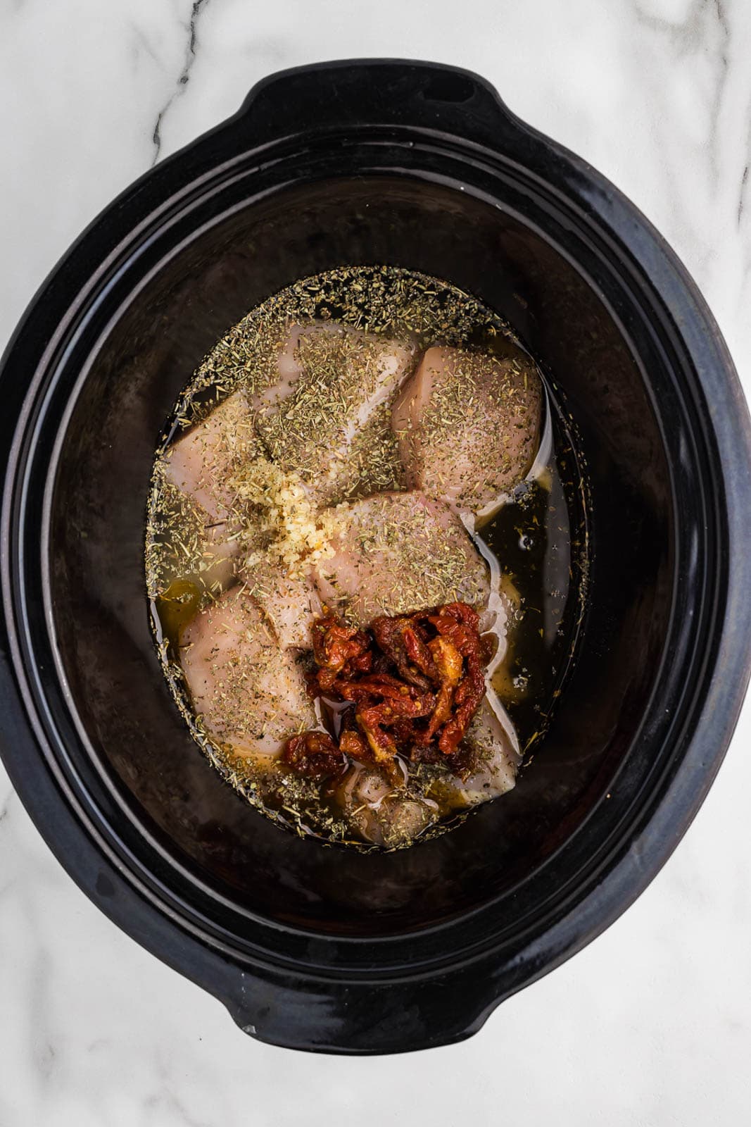 overhead shot of black slow cooker with chicken breasts in broth, garlic, seasoning and tomatoes