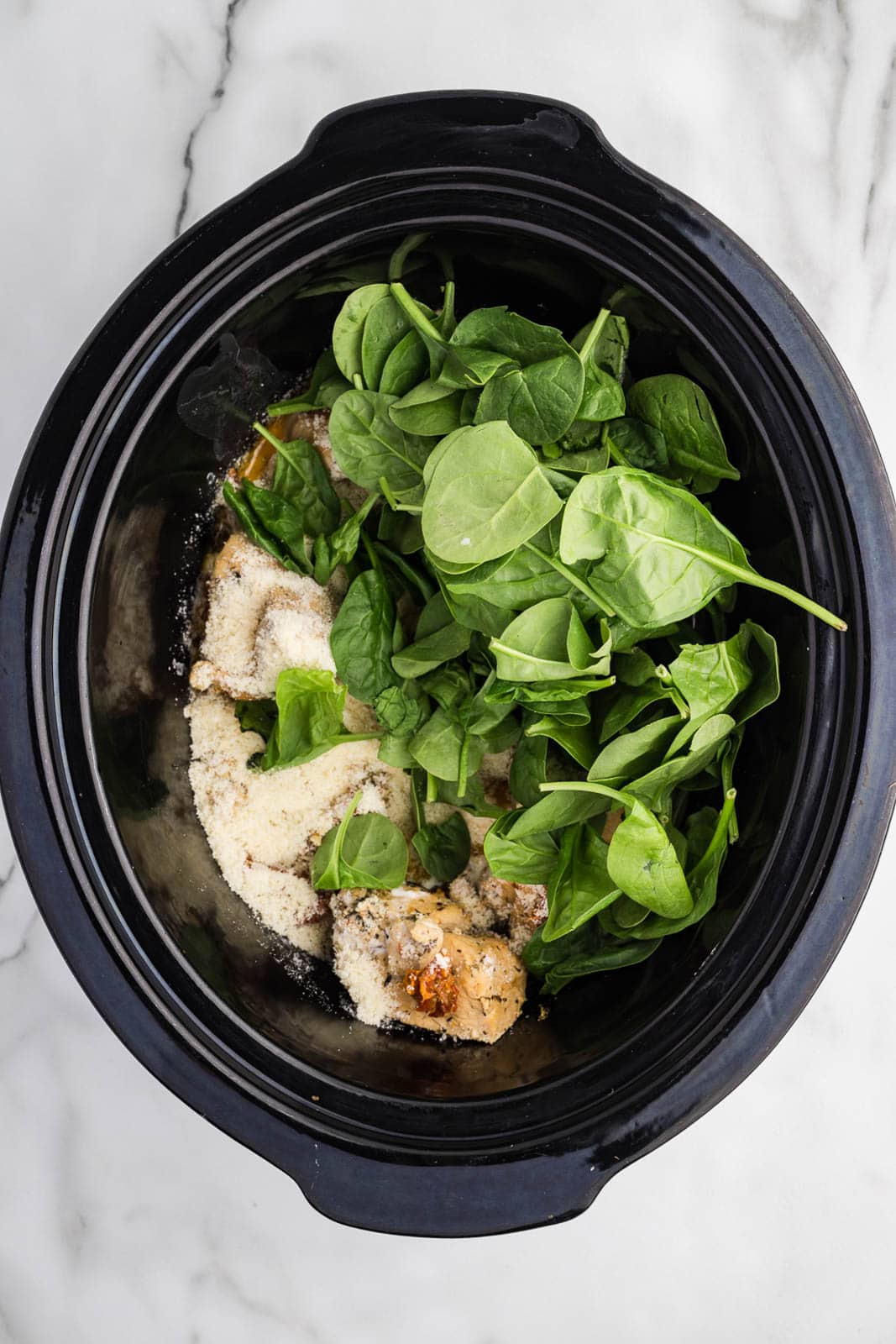 black slow cooker with cooked chicken and addition of raw spinach leaves