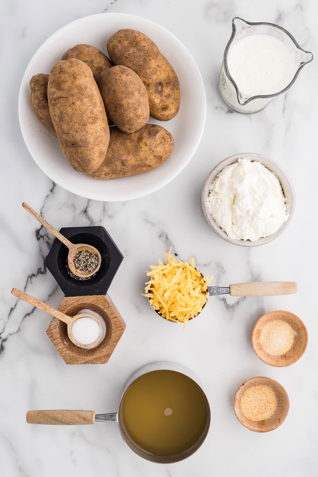 shot of ingredients to include white bowl of russet potatoes, measuring spoons with salt, pepper, measuring cup of chicken broth, cup of cheese, bowl of cream cheese, small bowls of garlic powder and onion powder