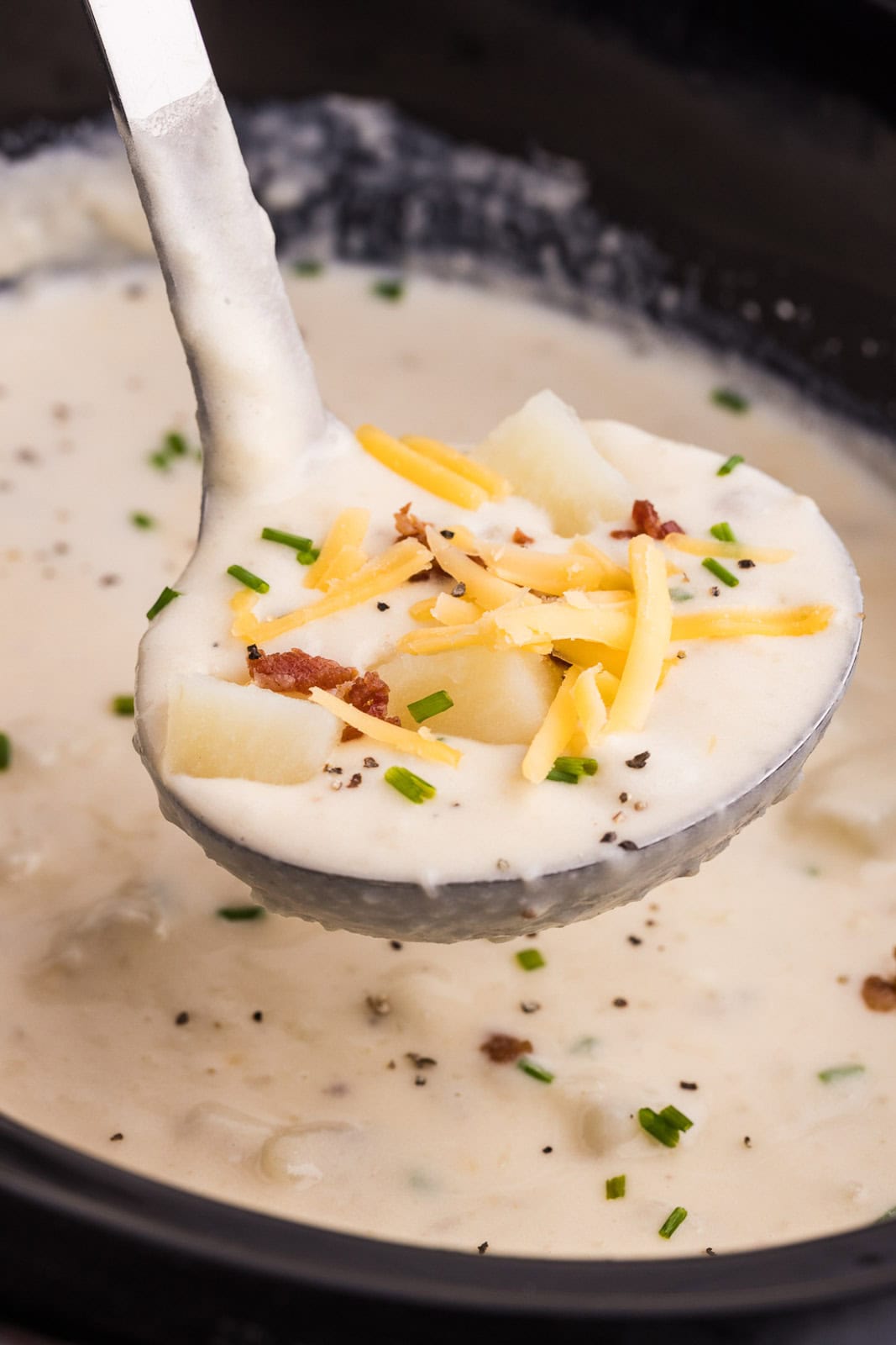 closeup of ladle containing potato soup over black slow cooker with soup