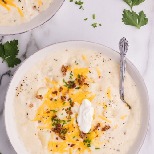 overhead shot of white bowl with silver spoon containing potato soup garnished with sour cream cheddar cheese, back and green onion, with second bowl in background surrounded by parsley