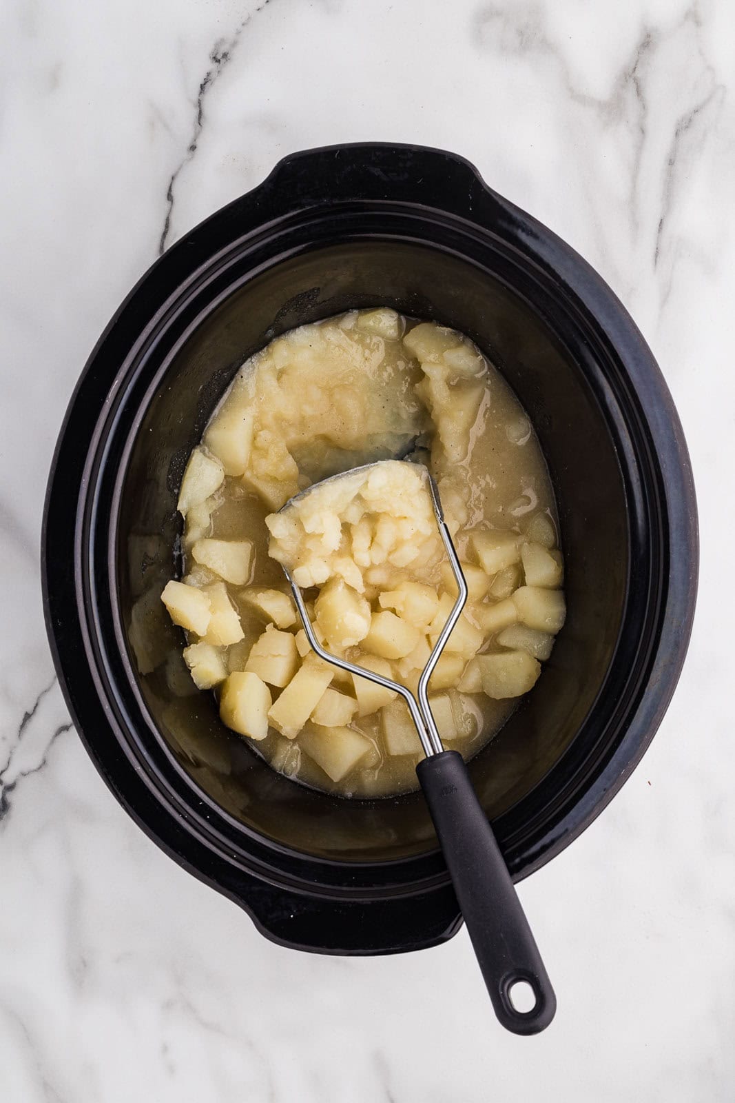 cooked potatoes in slow cooker with potatoes being mashed with masher