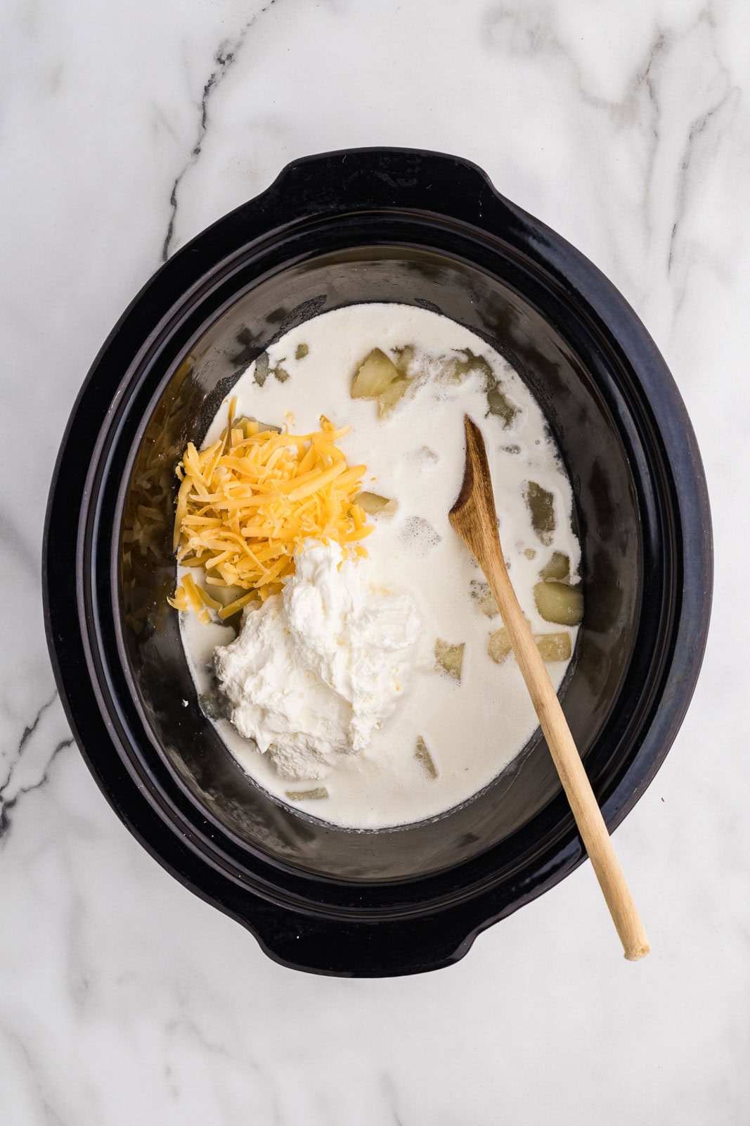 black slow cooker with wooden spoon, stirring in cream cheese, cheddar cheese and cream