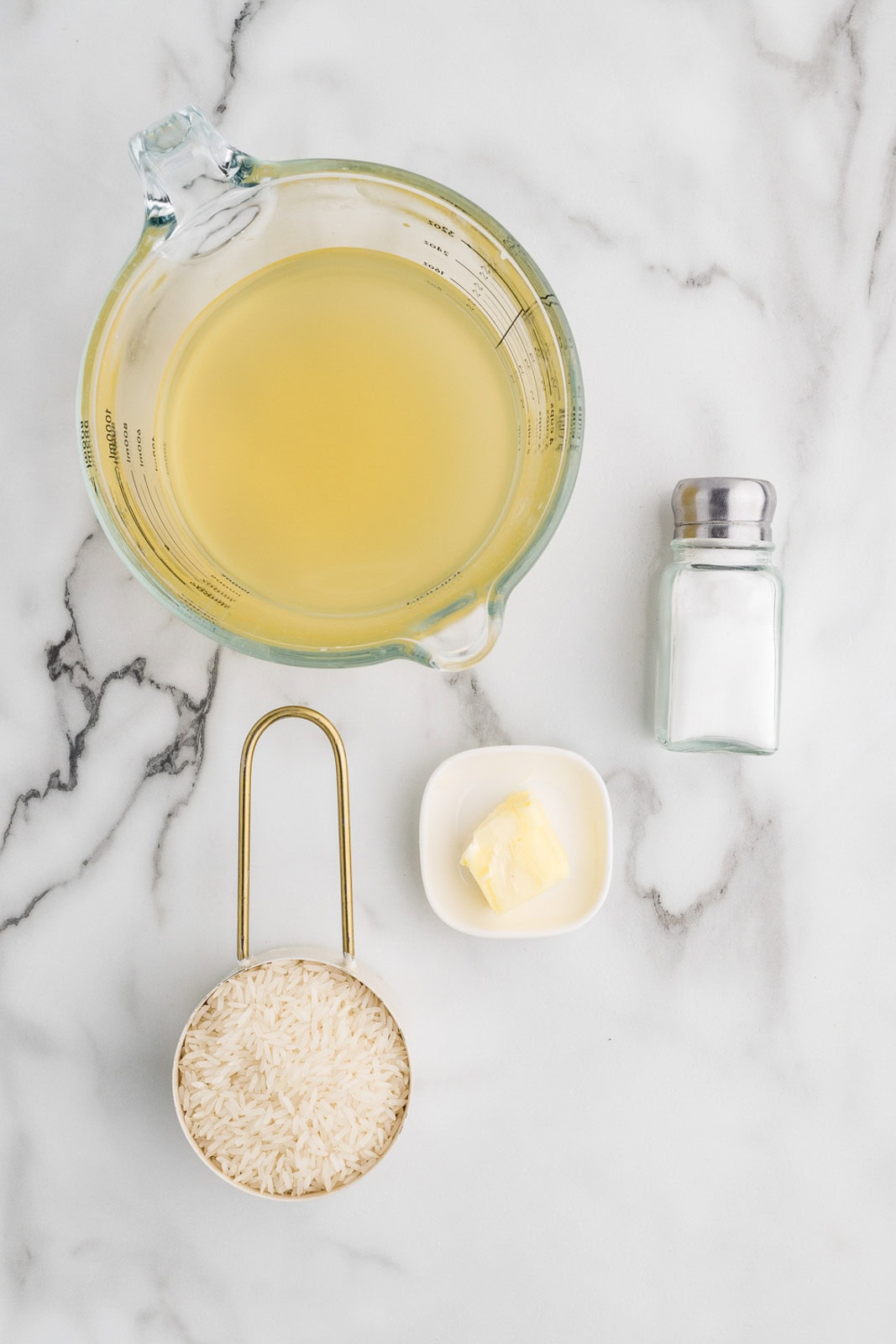 slow cooker rice ingredients to include glass measuring cup of broth, salt shaker, measuring cup of rice, and small bowl of butter