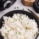black bowl of cooked rice with cilantro, surrounded by crockpot, salt and pepper containers with spoons, chopsticks, sprig of cilantro and black and white dish towel