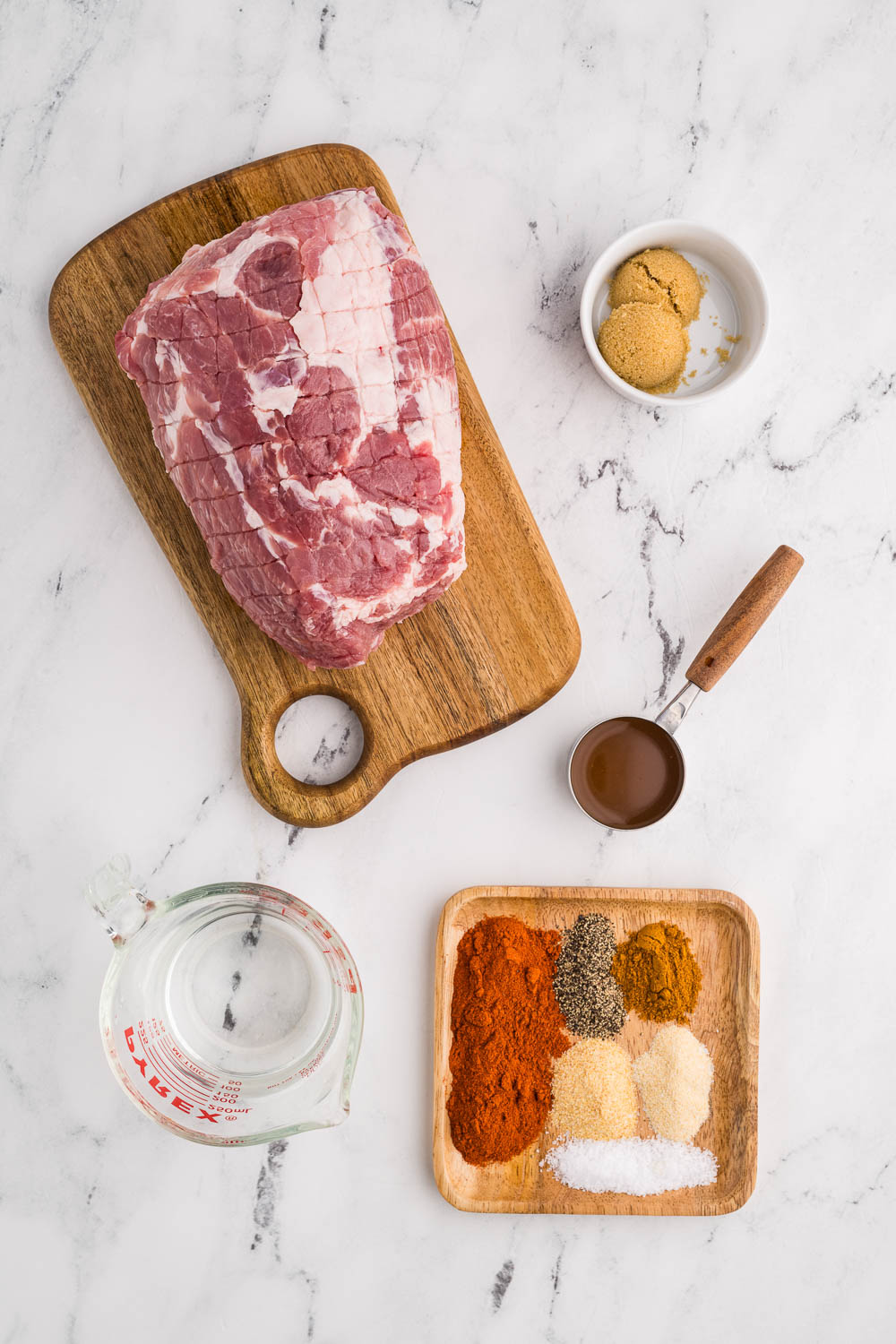 ingredients to include pork shoulder on wooden cutting board surrounded by white bowl of brown sugar, measuring cup with vinegar, glass measuring cup of water, and wooden tray with paprika, pepper, cayenne pepper, garlic and onion powder, and kosher salt