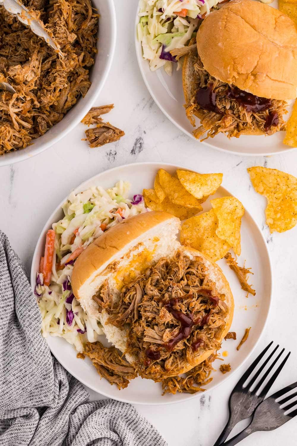 overhead view of white plate with open faced bun with pulled pork, Cole slaw and chips, with another white plate containing the same in background, with white bowl of pulled pork, two black forks and gray towel