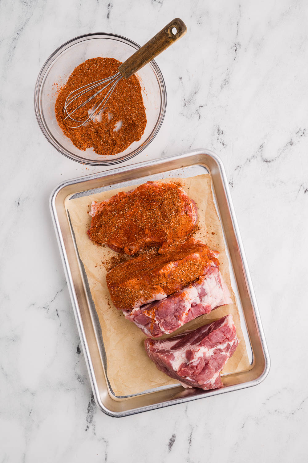 sheet pan lined with parchment paper with half covered pork with seasoning, with seasoning rub in clear bowl and whisk in background