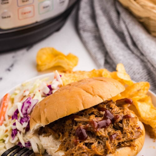 side shot of white plate with pulled pork on bun with Cole slaw and chips, with instant pot and basket of buns in background on gray towel
