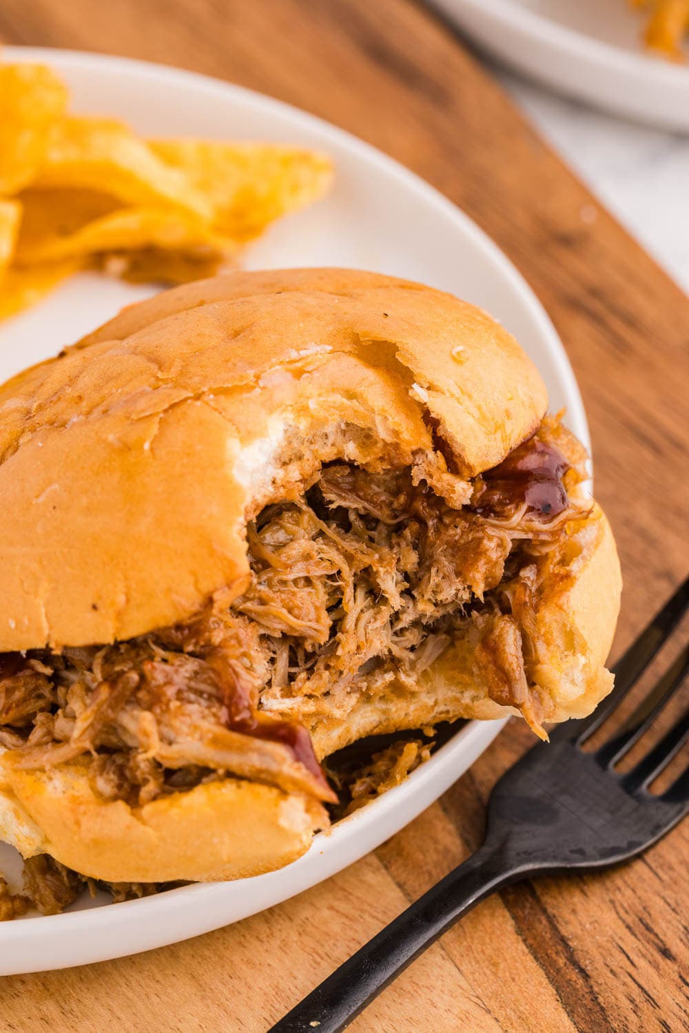white plate with pulled pork on bun with chips, black fork in front and bite taken of sandwich