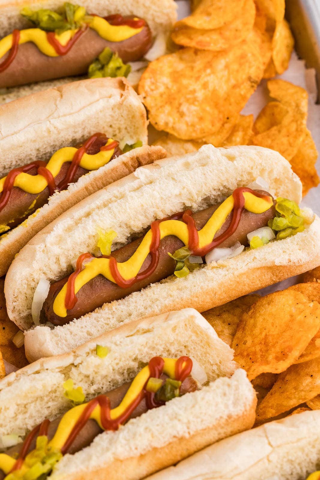closeup of 4 hot dogs in buns garnished with ketchup, mustard, relish, onions interspersed with bbq potato chips