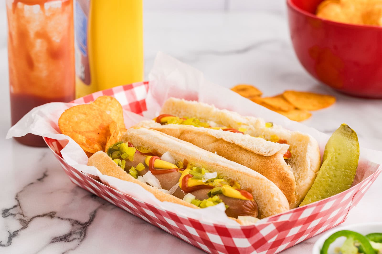 red and white paper tray with 2 dressed hot dogs, potato chips and dill pickle with bottle of ketchup and mustard, red bowl of potato chips in background