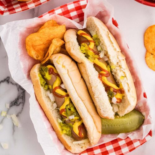 overhead view of red and white checked paper tray holding 2 dressed hotdogs, bbq potato chips and dill pickle surrounded by white bowl of onions, red bowl of potato chips and other trays with hot dogs