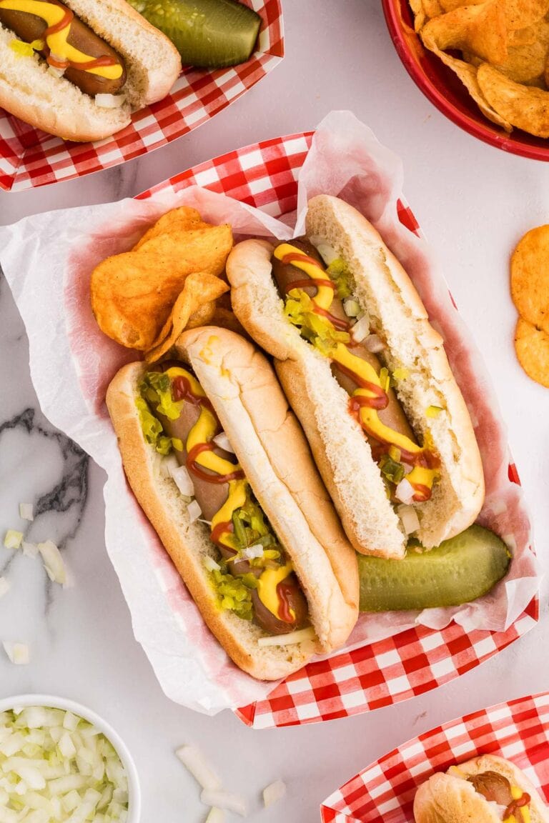 overhead view of red and white checked paper tray holding 2 dressed hotdogs, bbq potato chips and dill pickle surrounded by white bowl of onions, red bowl of potato chips and other trays with hot dogs