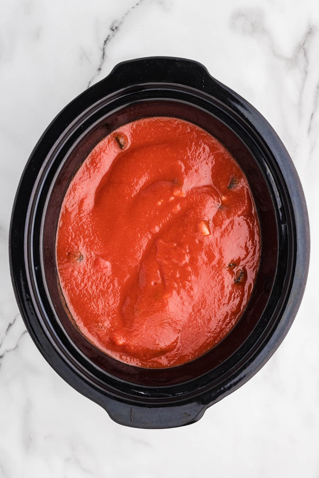 overhead view of chicken thighs and veggies, covered by tomato puree mixture in black slow cooker