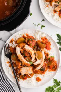 white dinner plate with chicken Cacciatore over a bed of rice and black dinner fork, with another finished plate and black slow cooker in the background