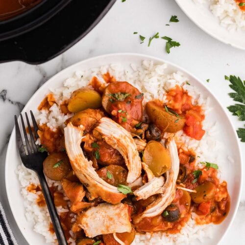 white dinner plate with chicken Cacciatore over a bed of rice and black dinner fork, with another finished plate and black slow cooker in the background