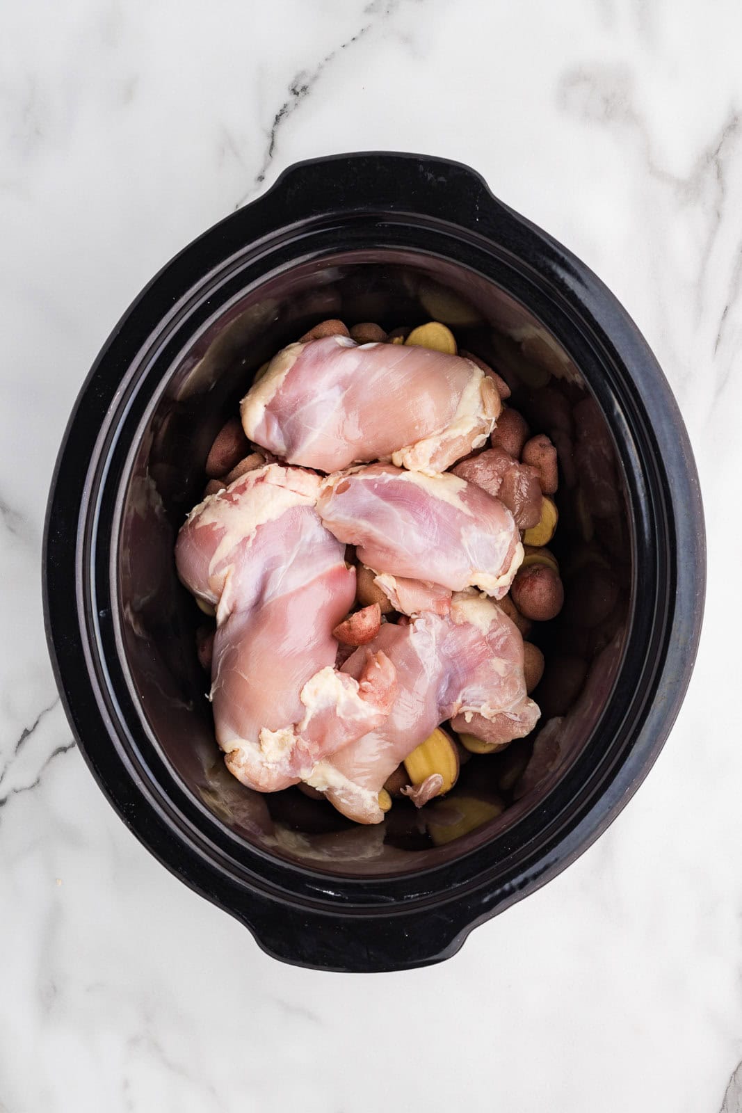 overhead view of black slow cooker with potatoes and chicken thighs on top