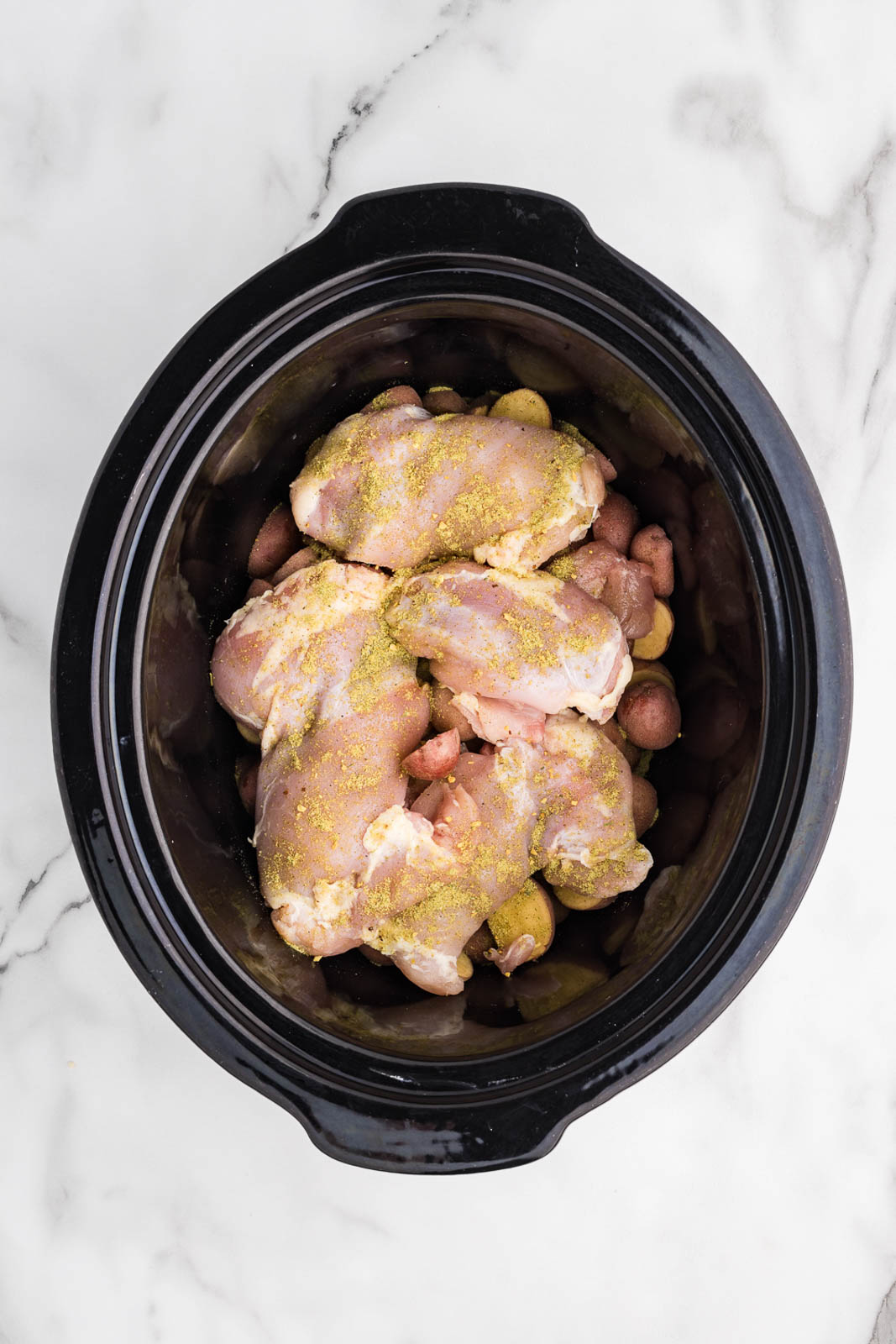 overhead view of black slow cooker with potatoes and chicken thighs on top, sprinkled with seasoning