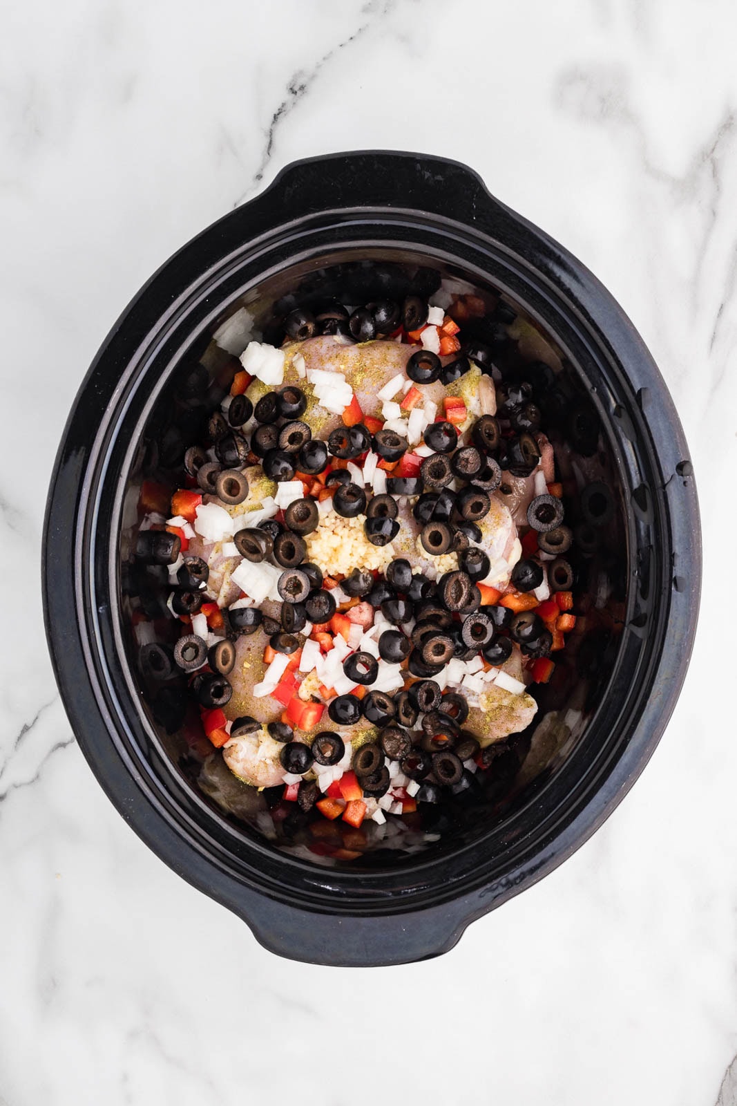 overhead view of black slow cooker with potatoes and chicken thighs with black olives, onions, garlic and red peppers
