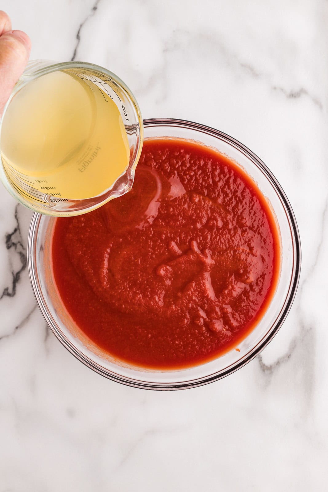 overhead view of glass bowl of tomato puree with glass measuring cup pouring chicken broth