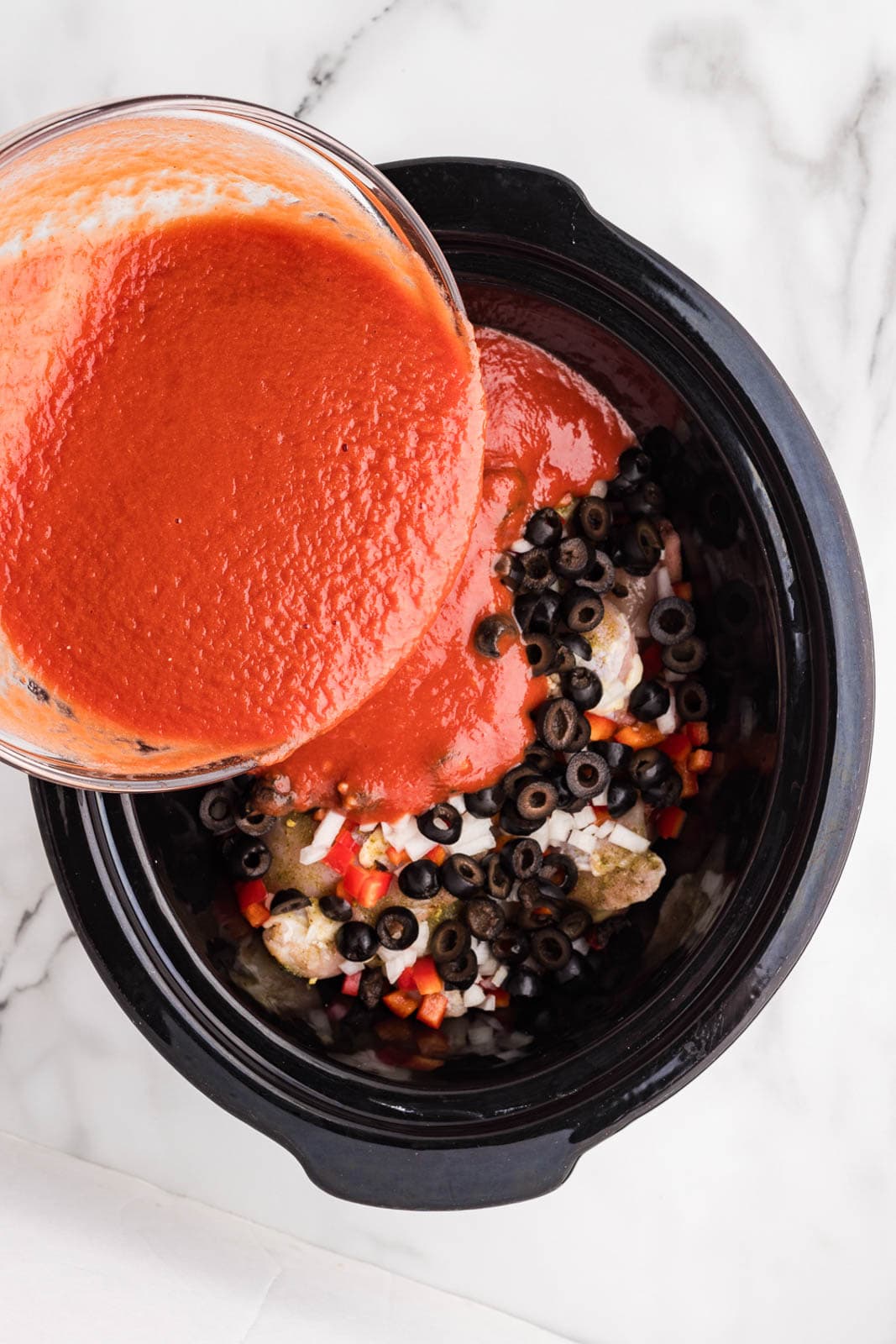 overhead view of black slow cooker, with chicken thighs and vegetable mixture, and glass bowl of tomato puree mixture being poured into slow cooker