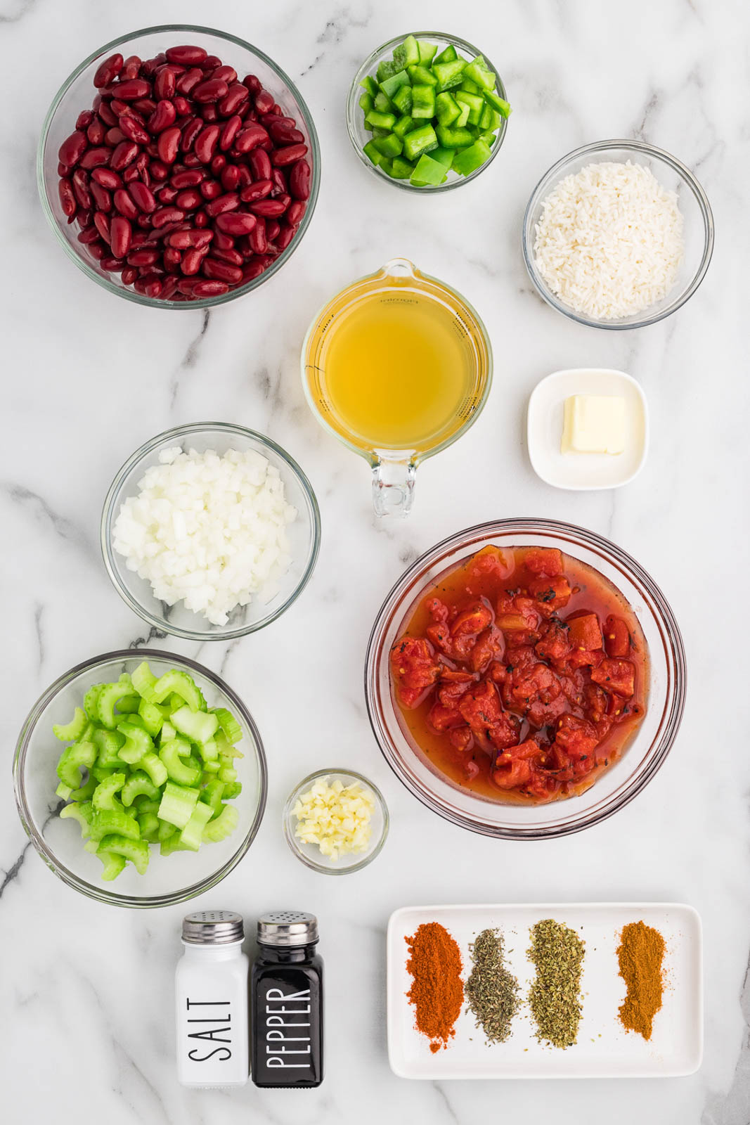 clear bowls containing ingredients to include red kidney beans, green pepper, fire roasted tomatoes, diced onion, diced celery, minced garlic, butter, salt and pepper shakers, and white plate with seasonings of paprika, thyme, oregano, cayenne pepper, white rice, and chicken broth