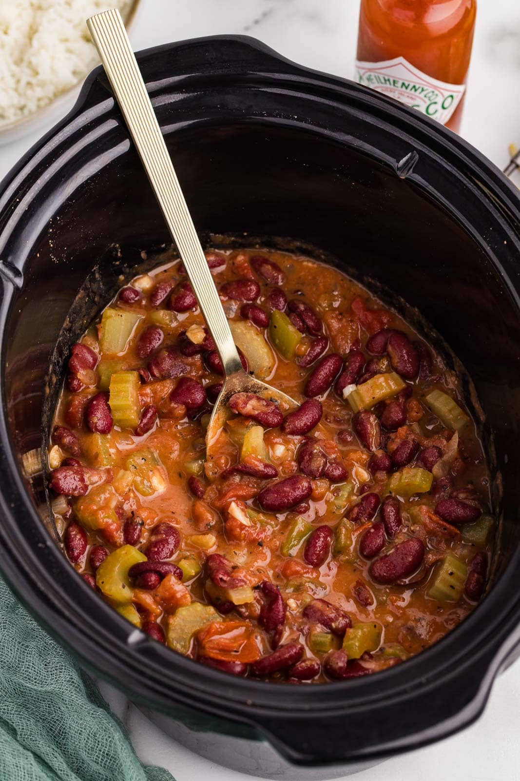 closeup of red bean mixture in black slow cooker with large gold ladle with hot sauce and bowl of white rice in background
