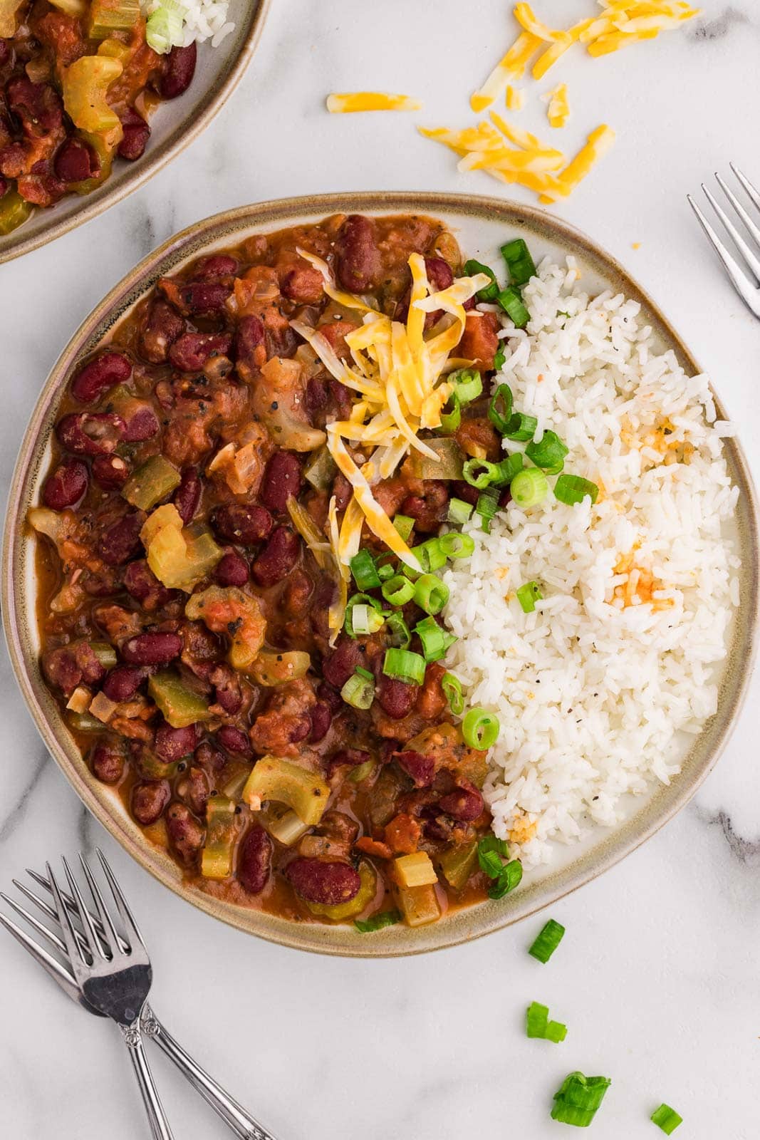 overhead view of plate of red beans and rice sprinkled with green onions, with 3 forks surrounding plate, and another dinner plate in background